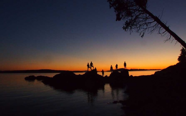 the silhouette of a group of people standing on rocks jutting out of a body of water is illuminated against the sunset.